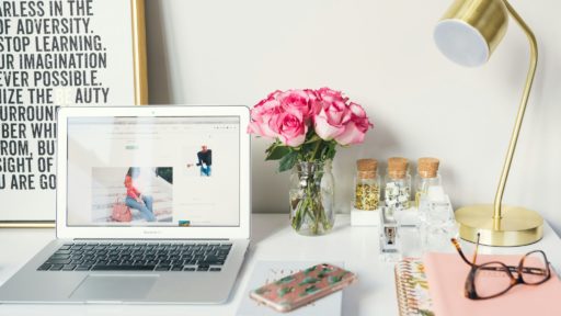 A photo of a clean white desk with a laptop sat on top of it. To the right side are some pink flowers in a small vase and a golden table lamp.